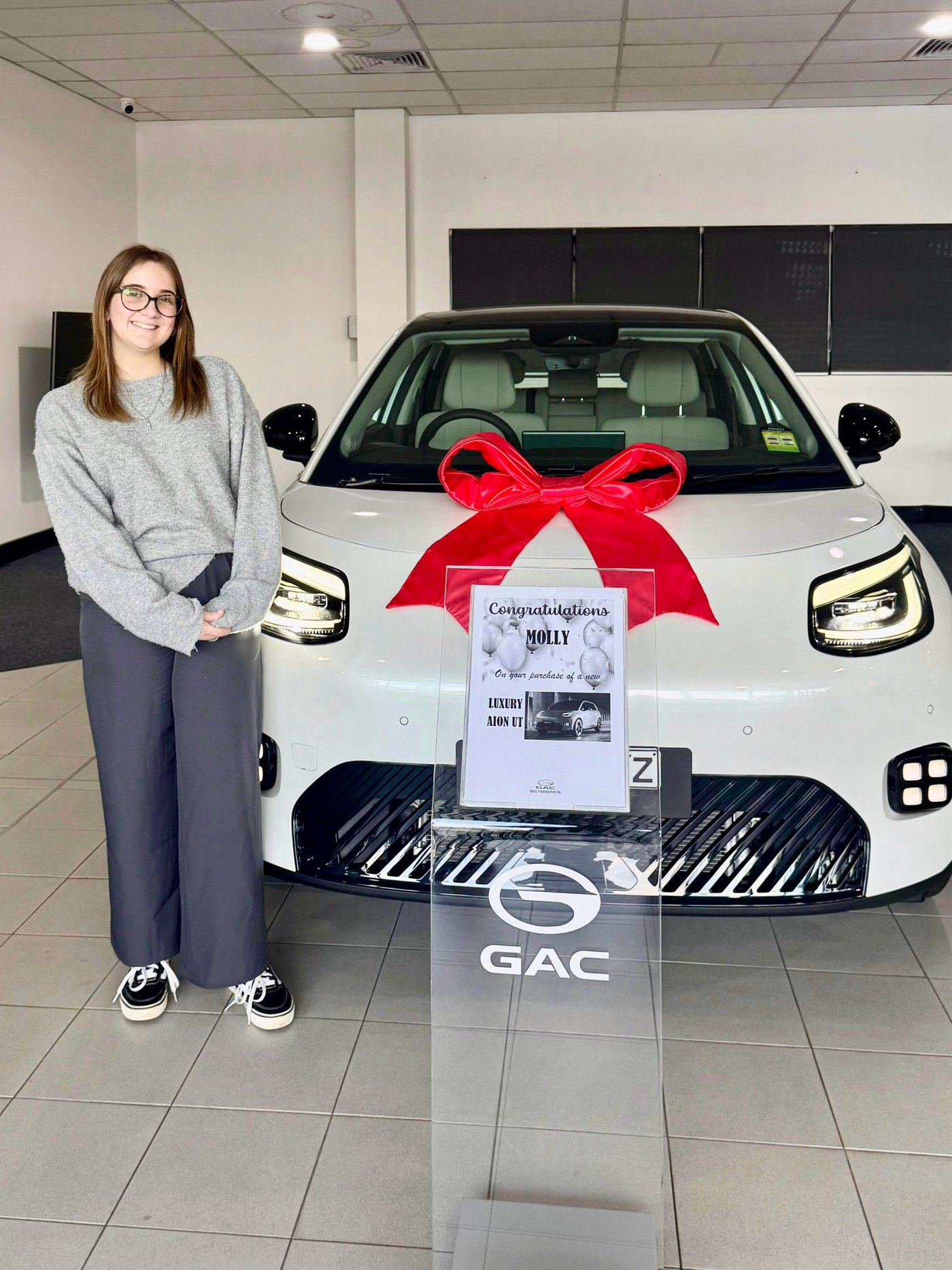 A customer stands beside a white AION UT with a red bow in the Aion Parramatta GAC showroom, next to a printed card reading Congratulations MOLLY on your purchase of a new LUXURY AION UT.